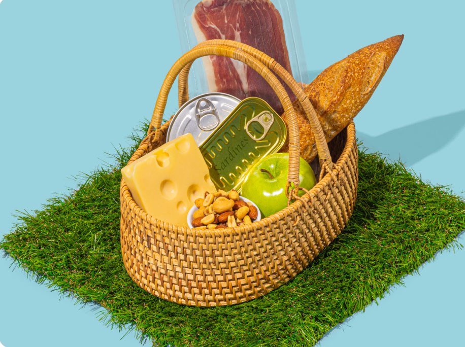 A basket with various foods, including cheese, nuts, apples, tinned seafood, cured meat and bread