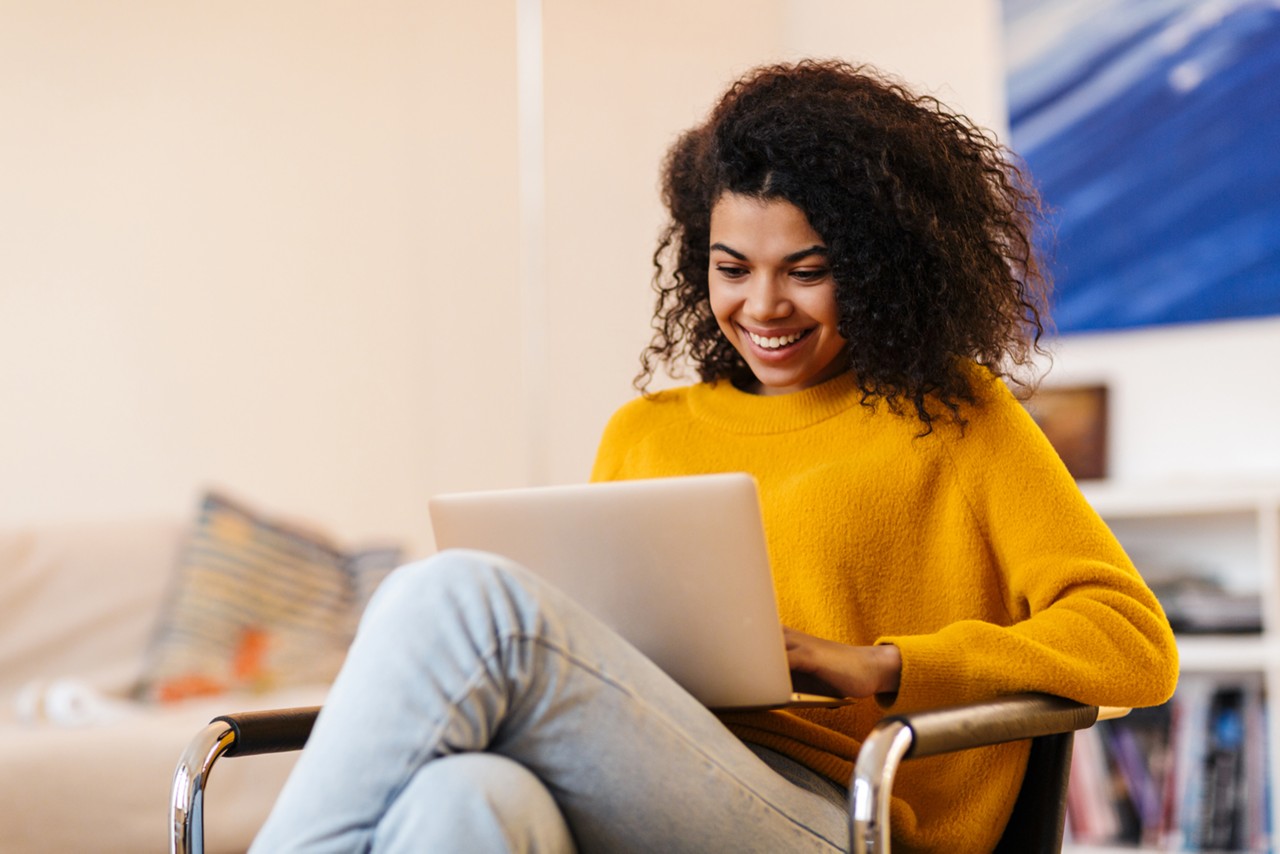 Image of cheerful african american woman using laptop while sitting on chair in living room; Shutterstock ID 1718299927; purchase_order: -; job: -; client: -; other: -