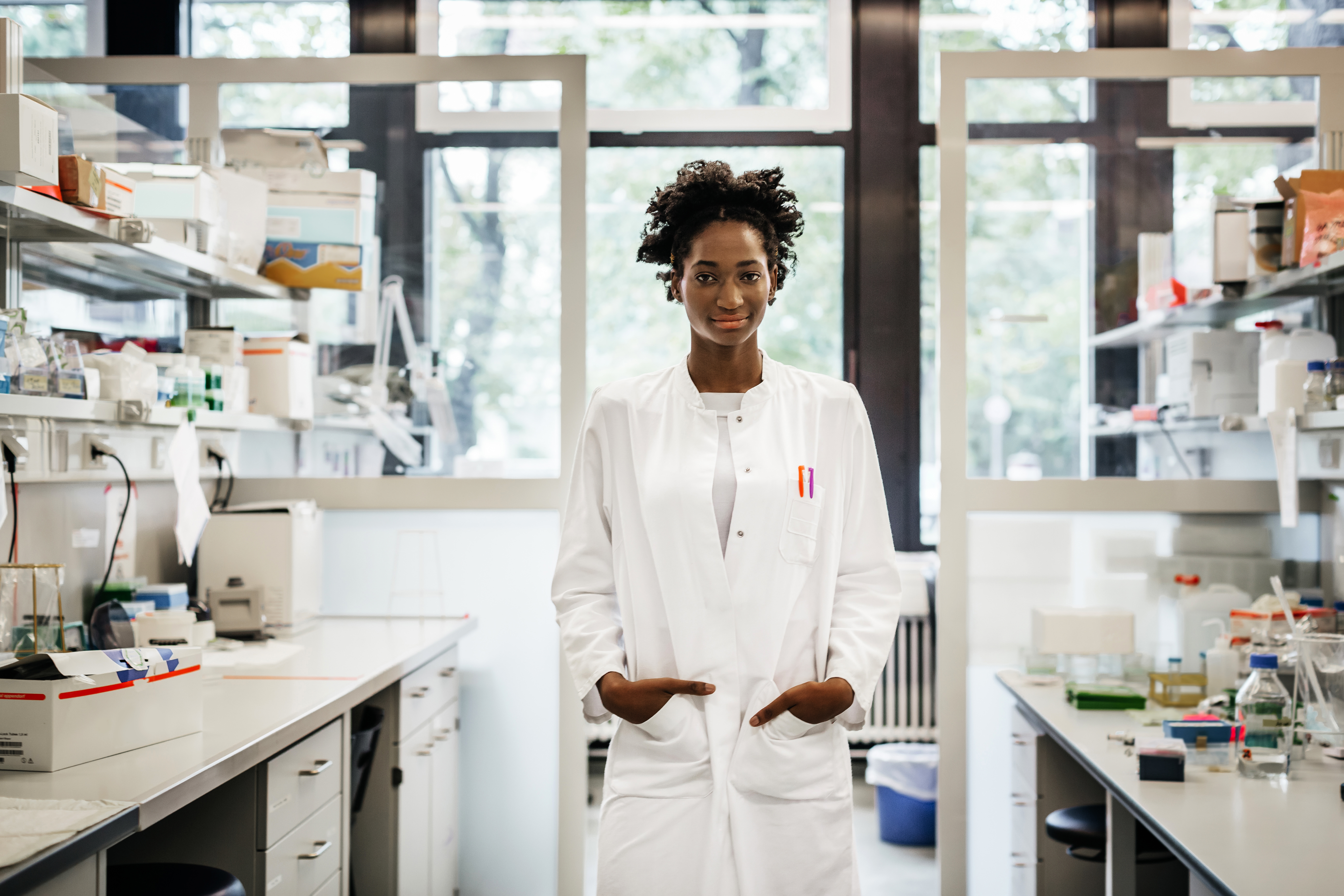 A black female scientist is standing in a laboratory between desks with scientific equipment and files. She's smiling into the camera with he hands in her coat pockets.