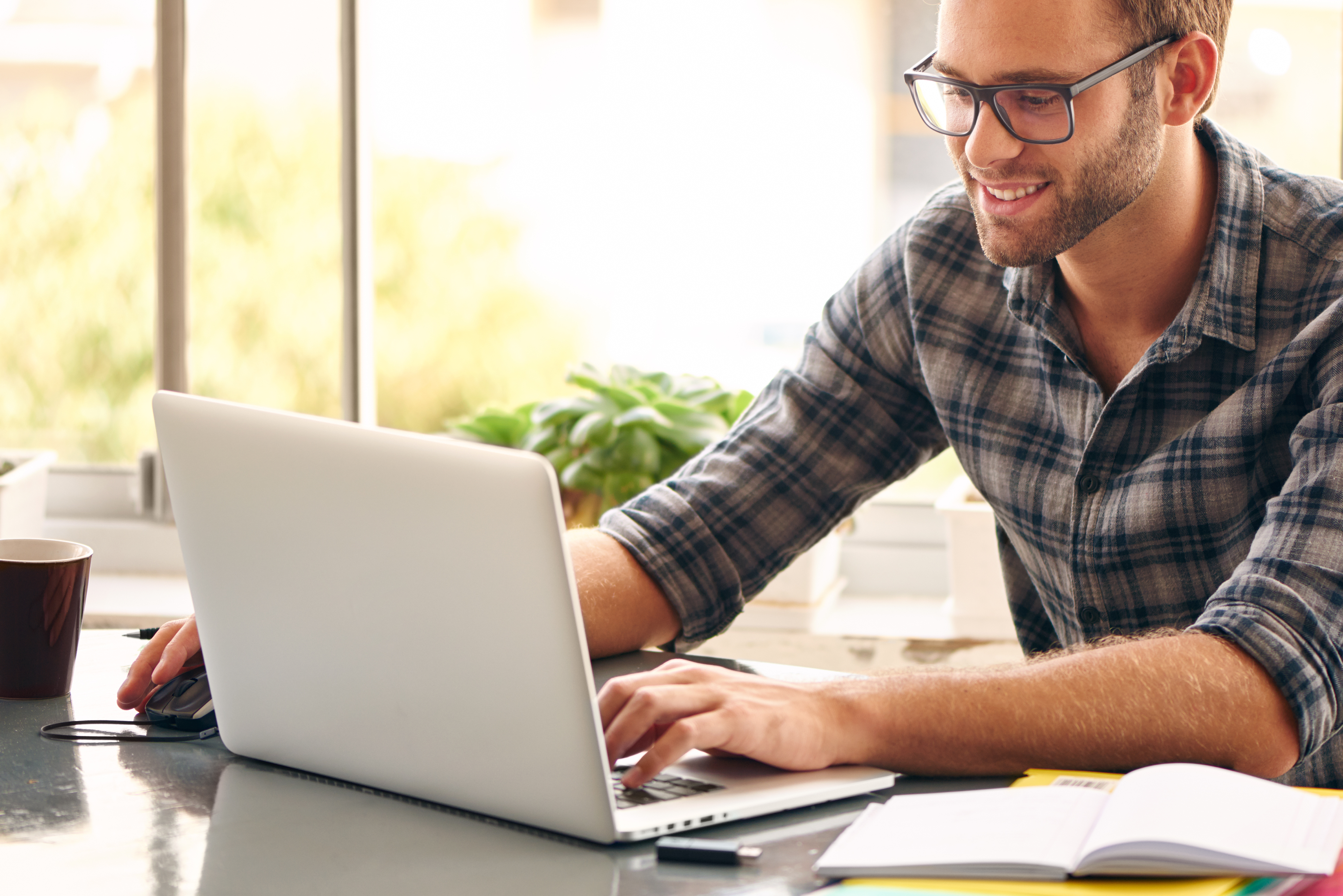 Happy young man, wearing glasses and smiling, as he works on his laptop to get all his business done early in the morning with his cup of coffee